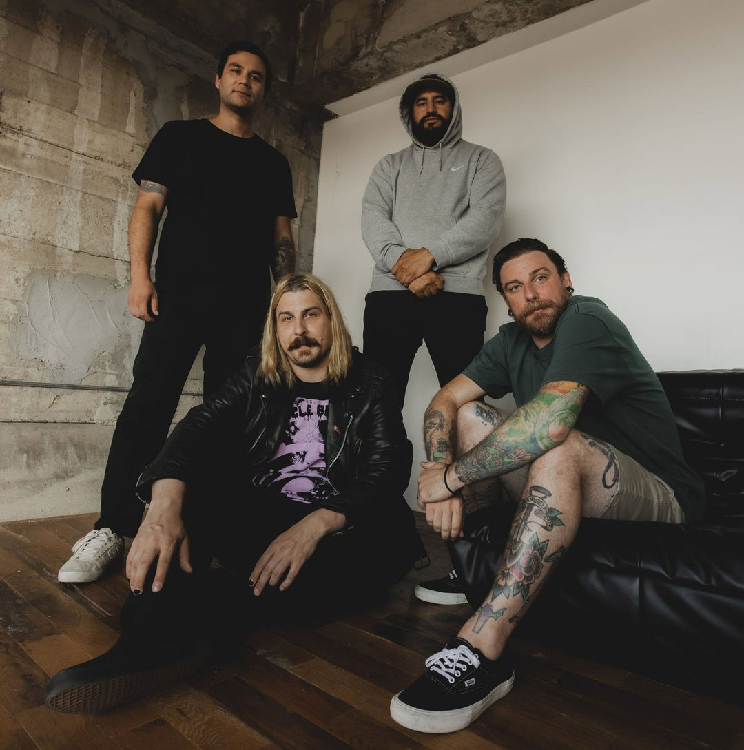 Four men pose indoors against a white and concrete wall; two are standing, two are sitting, one on the floor and one on a black couch. They have casual clothing and visible tattoos.