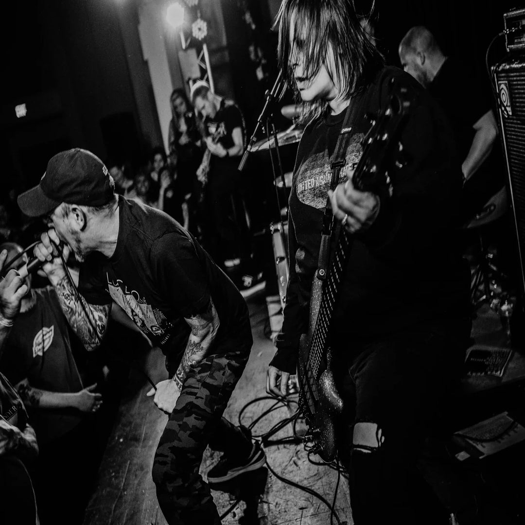 A black-and-white photo of a hardcore punk band performing on stage. The vocalist screams into the mic while the bassist plays intensely. The crowd watches closely in a dimly lit venue.