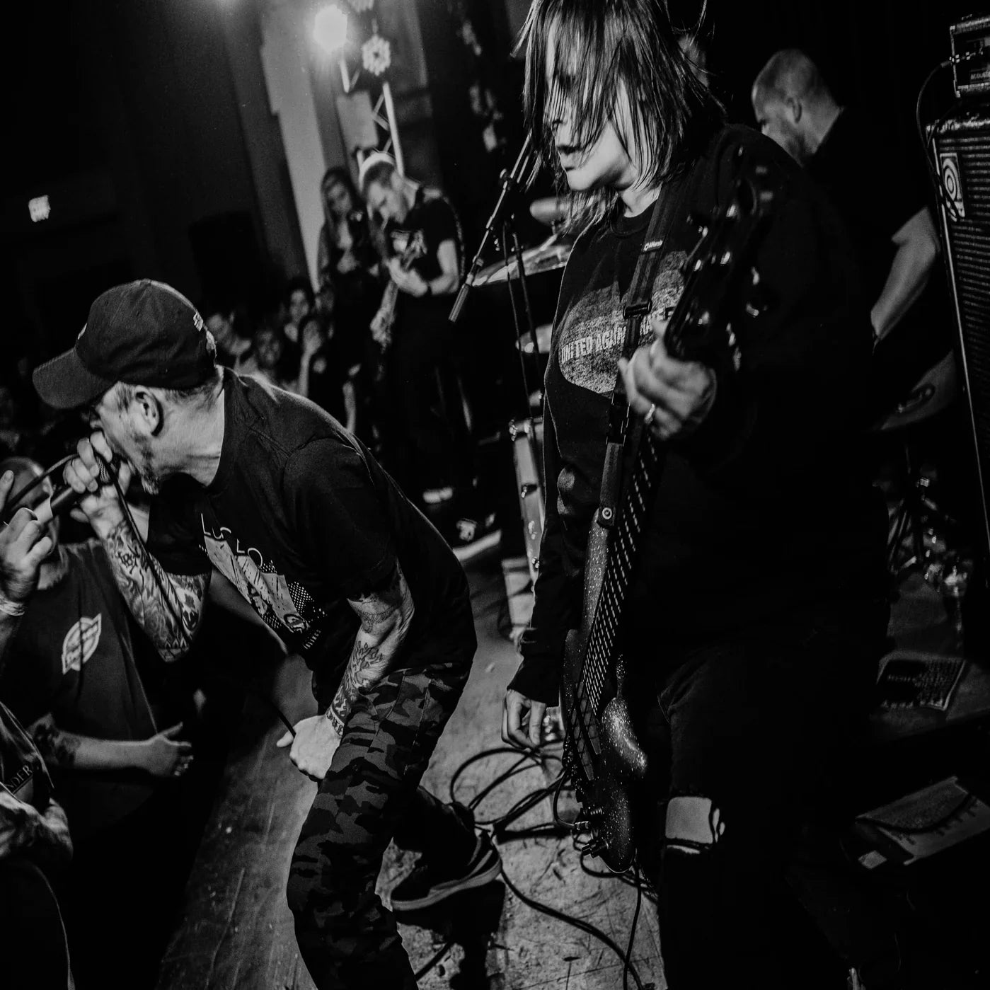 A black-and-white photo of a hardcore punk band performing on stage. The vocalist screams into the mic while the bassist plays intensely. The crowd watches closely in a dimly lit venue.