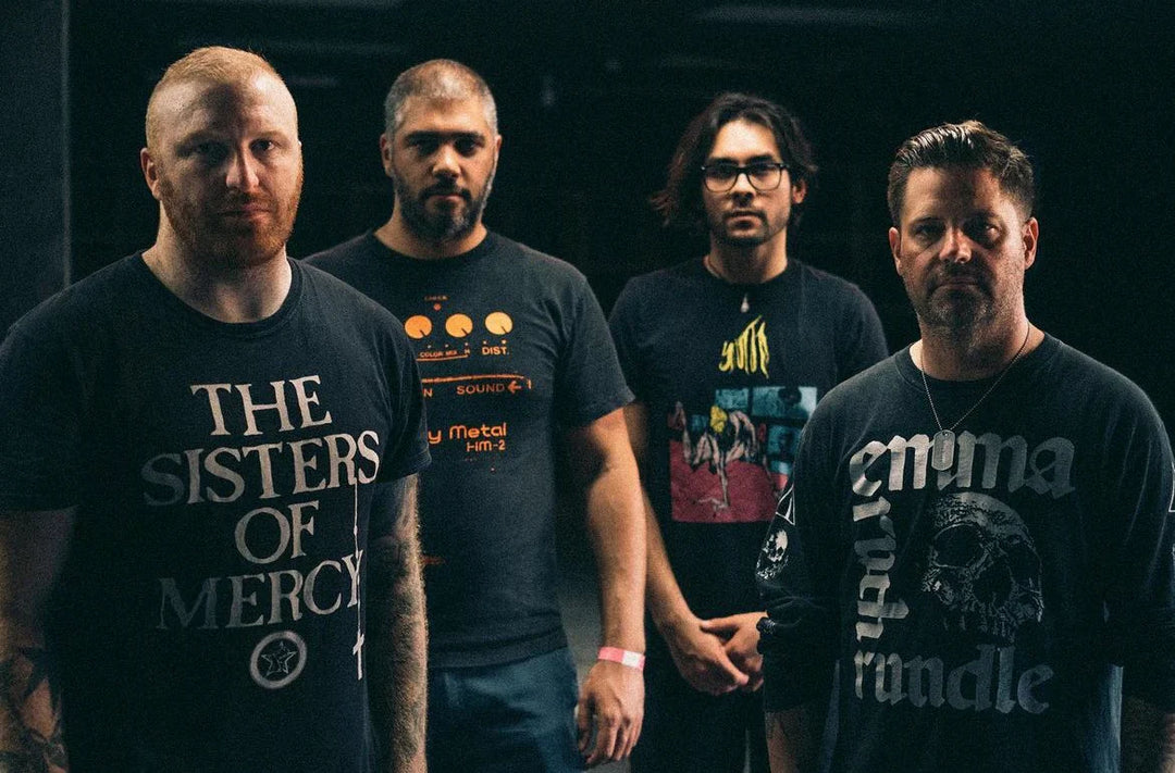 Four men stand together indoors, facing the camera. They wear dark band t-shirts and appear serious, with a dim, moody background behind them.