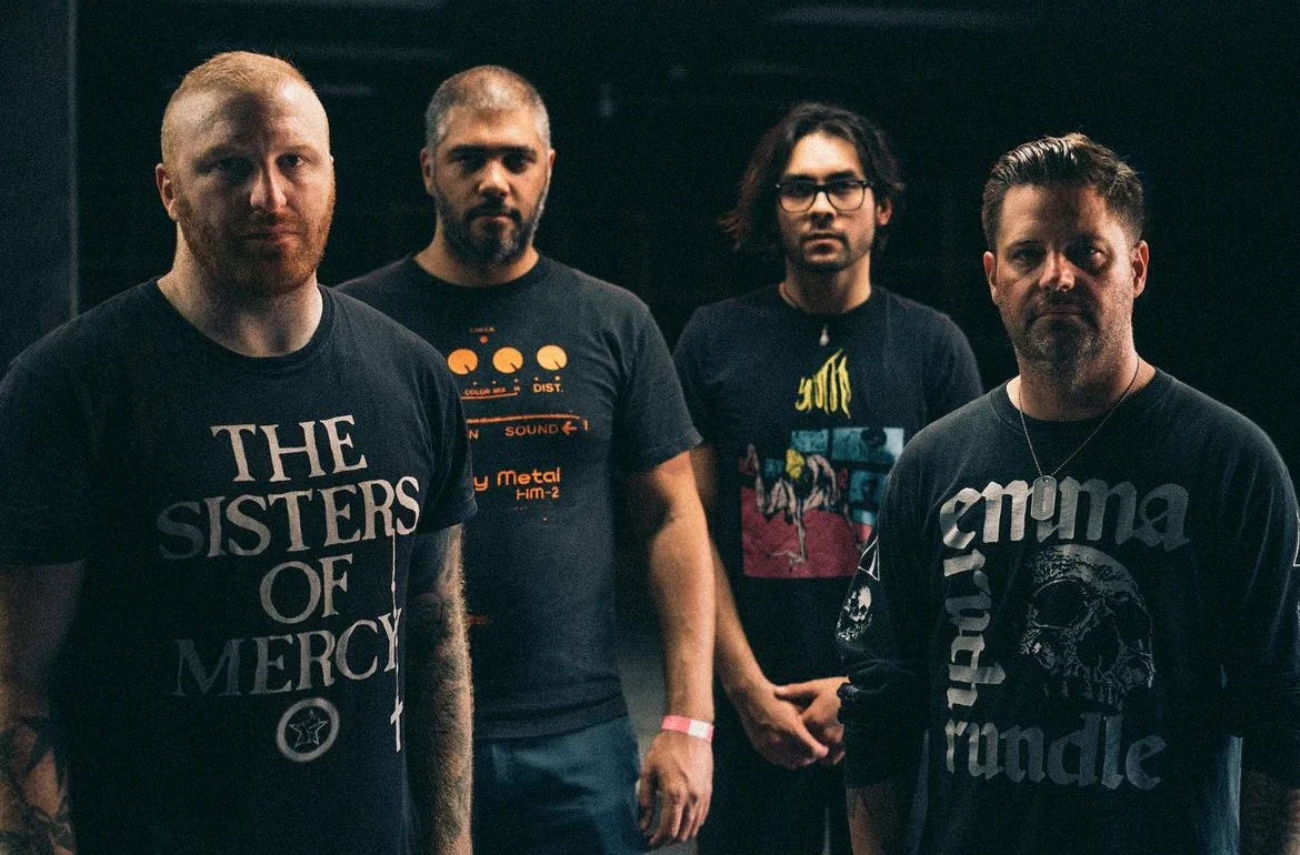 Four men stand together indoors, facing the camera. They wear dark band t-shirts and appear serious, with a dim, moody background behind them.