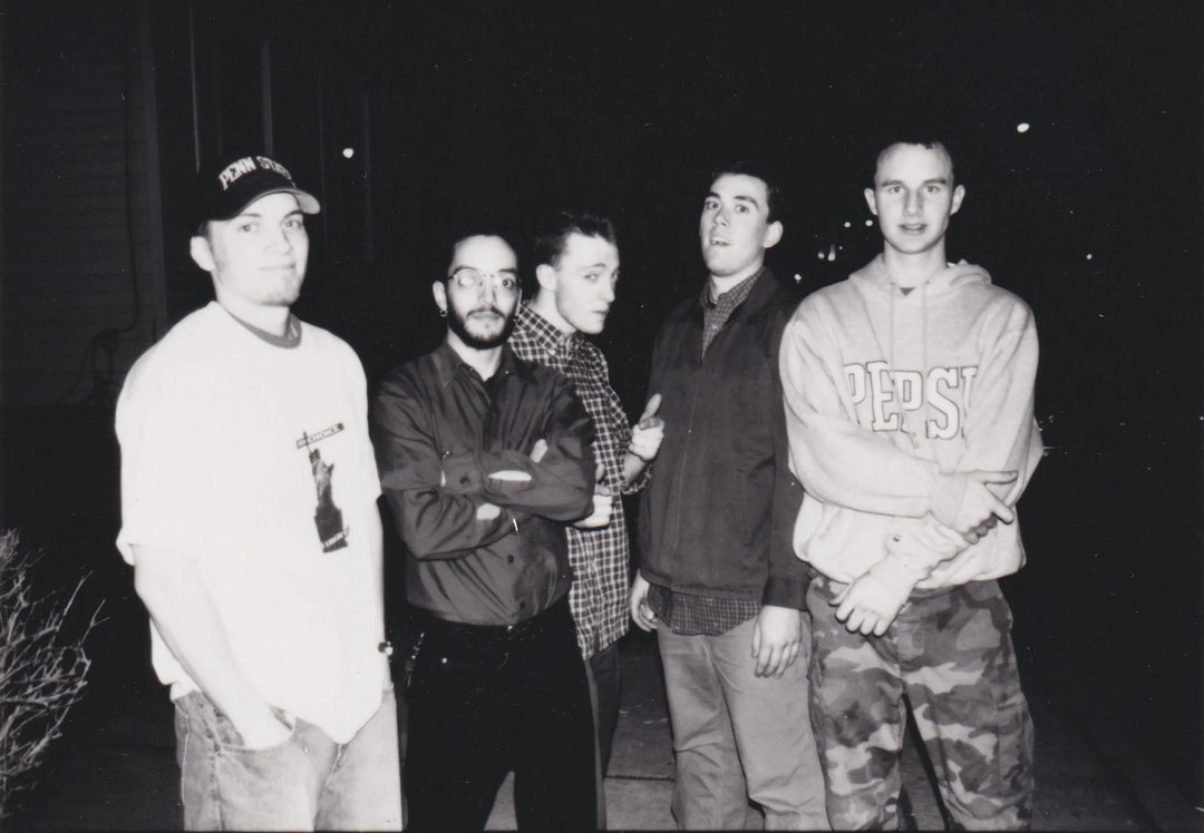 Five young men stand together outside at night. Four face the camera, one looks away. They wear casual clothes, including a Pepsi hoodie, cap, glasses, and plaid shirts. The scene is in black and white.