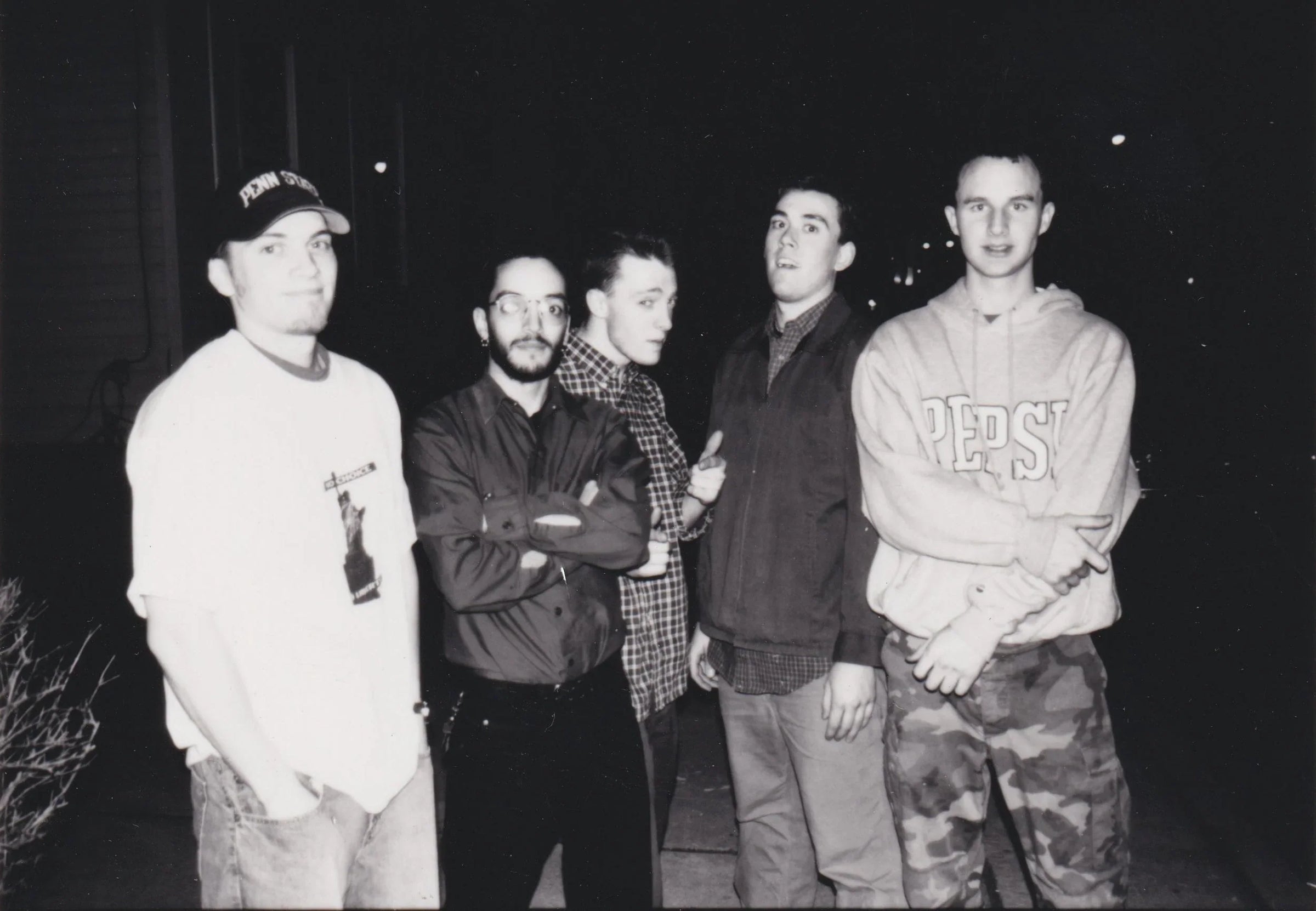 Five young men stand together outside at night. Four face the camera, one looks away. They wear casual clothes, including a Pepsi hoodie, cap, glasses, and plaid shirts. The scene is in black and white.