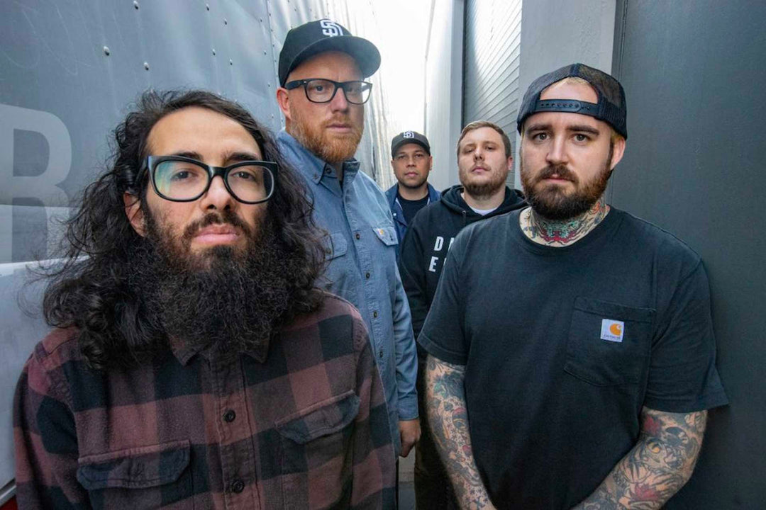 Five men stand in a narrow alleyway, some with serious expressions. One has a long beard and glasses, another has tattoos and a cap, and the rest wear casual clothes and hats.