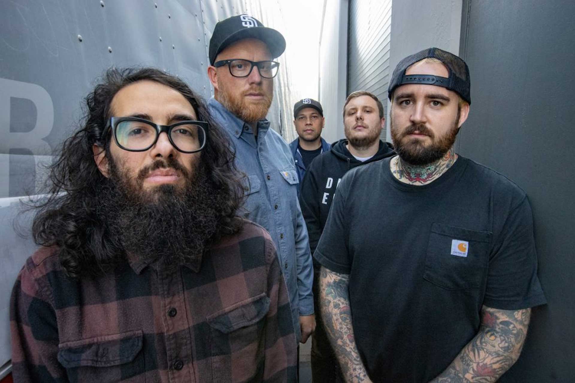 Five men stand in a narrow alleyway, some with serious expressions. One has a long beard and glasses, another has tattoos and a cap, and the rest wear casual clothes and hats.