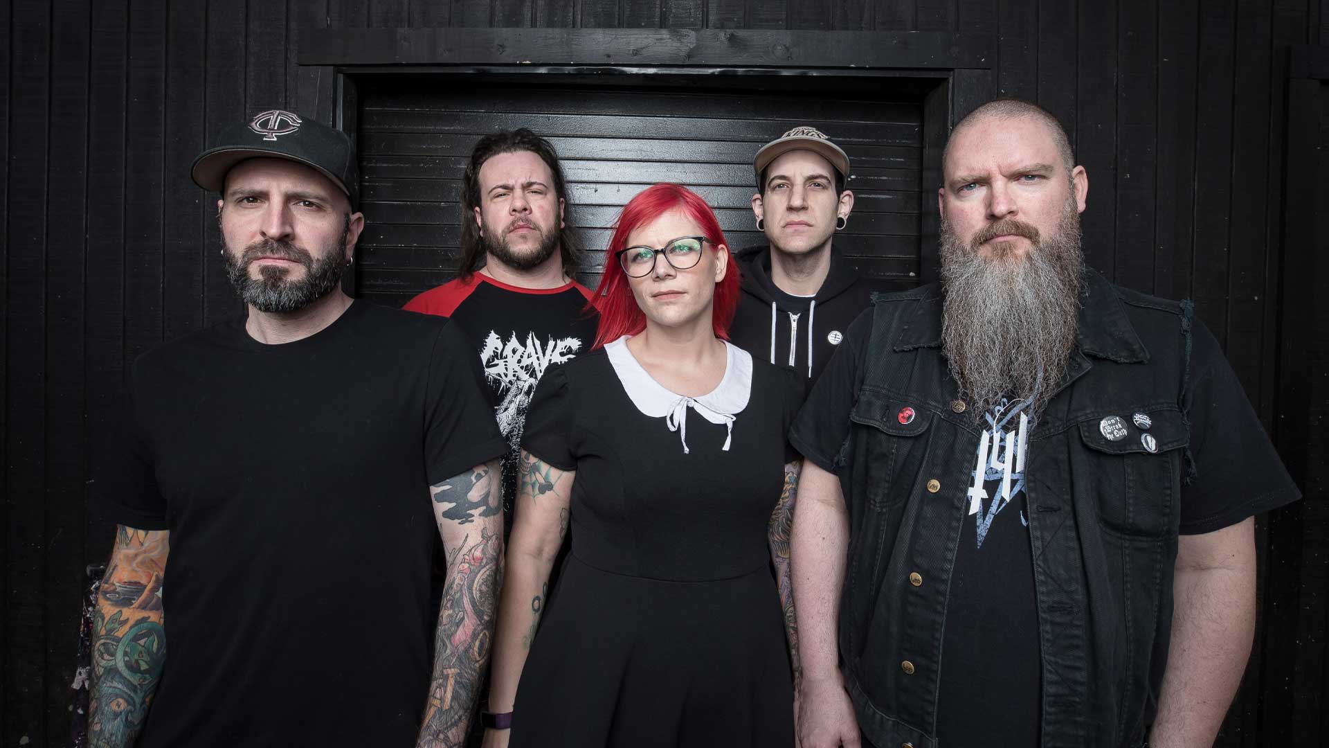 Five people stand in front of a dark wall, looking serious. The group includes three men with beards, one man in a cap, and a woman with red hair and glasses wearing a black dress with a white collar.
