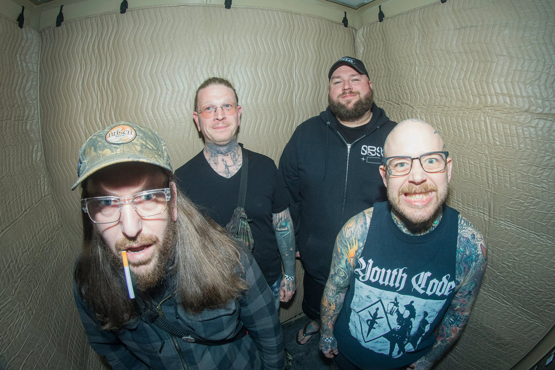 Four men with tattoos stand in a small room with padded walls. One wears a cap and glasses with a cigarette in his mouth, another wears a Youth Code shirt, and all are smiling or making faces at the camera.