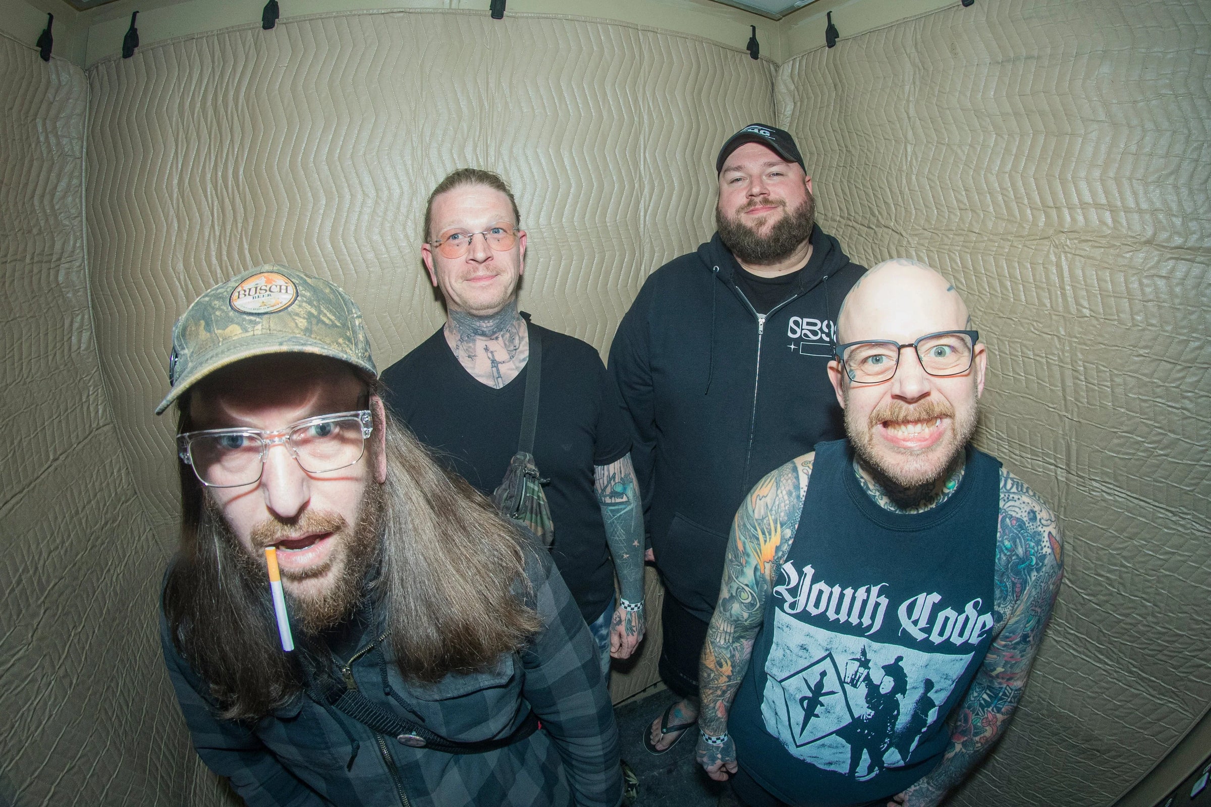 Four men with tattoos stand in a small room with padded walls. One wears a cap and glasses with a cigarette in his mouth, another wears a Youth Code shirt, and all are smiling or making faces at the camera.