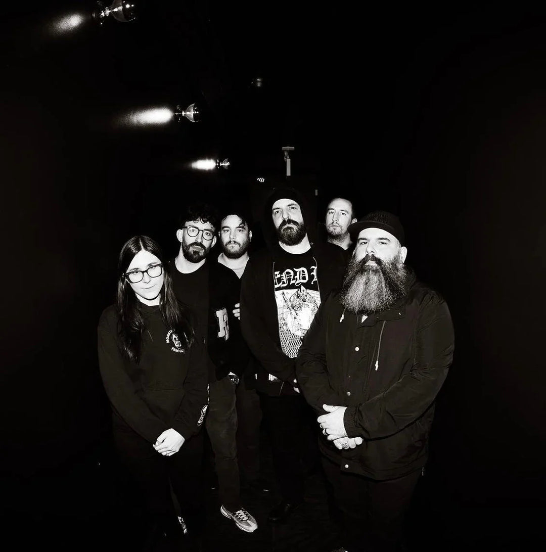 A group of six people, five men and one woman, stand together in a dark hallway, facing the camera. They are wearing dark clothing, and one man has a large beard. The image is in black and white.