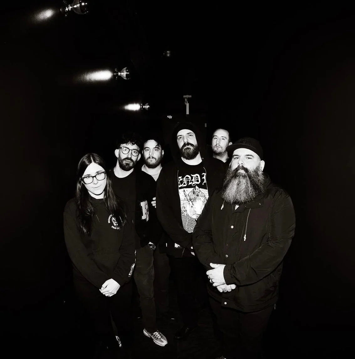 A group of six people, five men and one woman, stand together in a dark hallway, facing the camera. They are wearing dark clothing, and one man has a large beard. The image is in black and white.