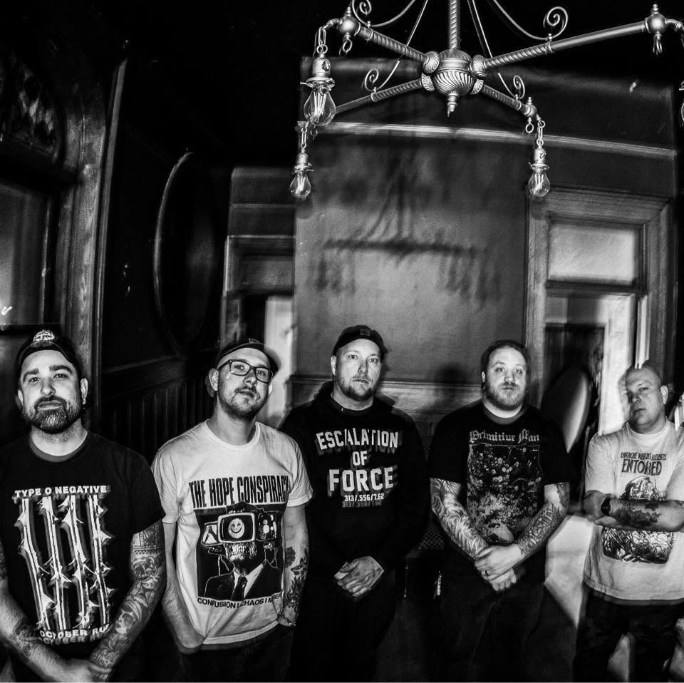 Five tattooed men stand indoors under a chandelier, facing the camera. They wear black graphic T-shirts and hats. The photo is in black and white, creating a moody, dramatic atmosphere.